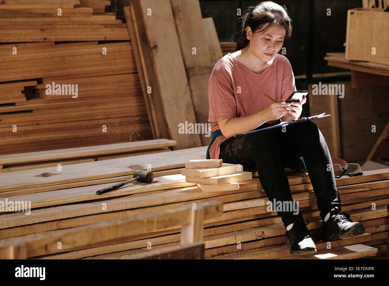 Asian female carpenter with clipboard on lap using smartphone while sitting on wooden planks in workshop Stock Photo