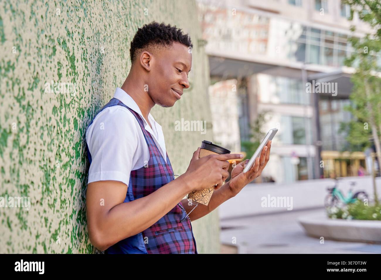Black man holding takeaway coffee and using tablet while standing and leaning against concrete wall in city during daytime Stock Photo