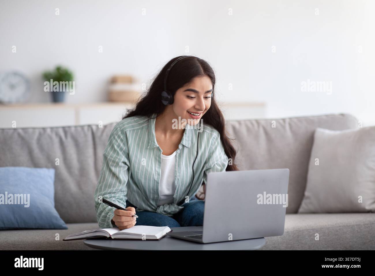 Indian female in headset writing down notes having online class on laptop sitting on sofa at home Stock Photo