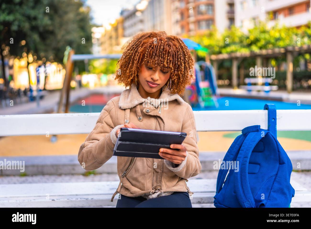 Mixed-race female teenager with blonde twists wearing brown jacket using tablet while sitting on bench with blue backpack in city park during daytime Stock Photo