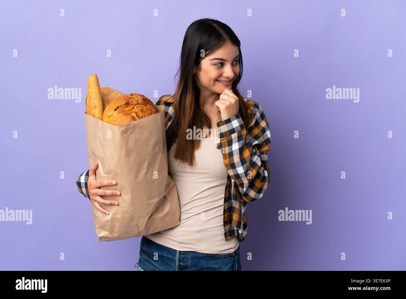 Hispanic woman carrying bag of bread looking at her side while standing against purple background in studio Stock Photo