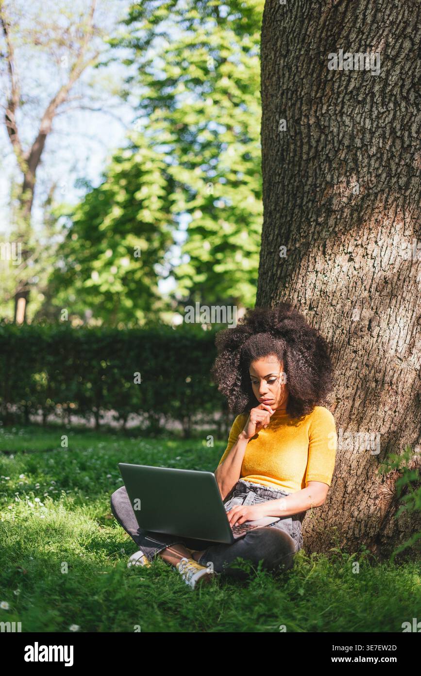 Hispanic female with curly hair leaning chin on hand working on laptop sitting against tree trunk in park during daytime Stock Photo