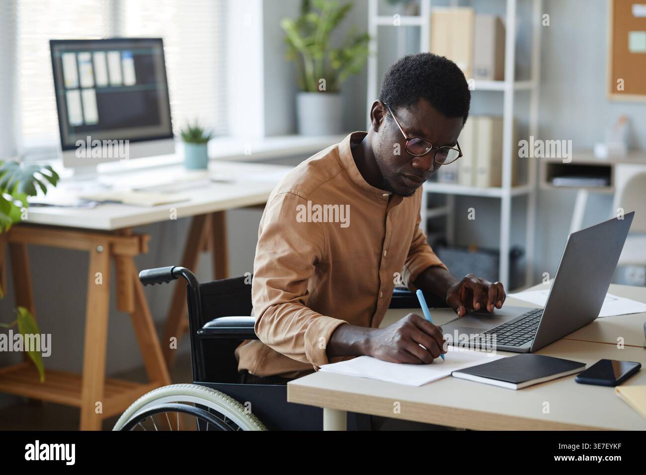 Black male disabled IT specialist writing on paper while working on laptop in office during daytime Stock Photo
