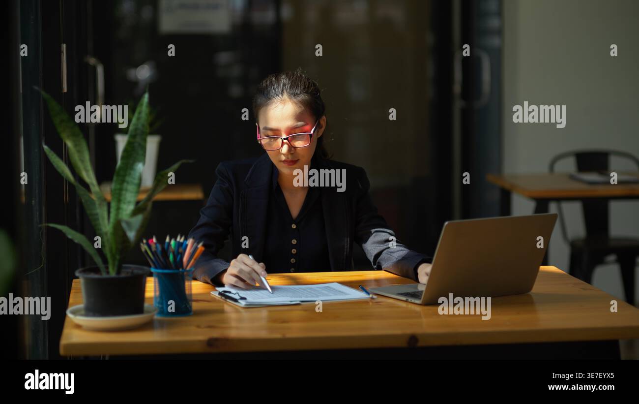 Asian woman checking documents while using laptop sitting at table in office Stock Photo