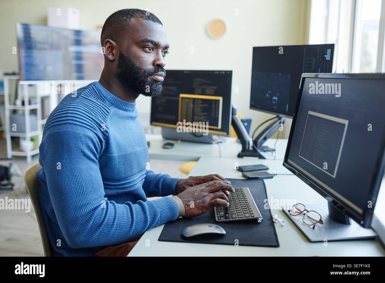 Black male programmer with beard typing codes on computer while sitting at table in office Stock Photo