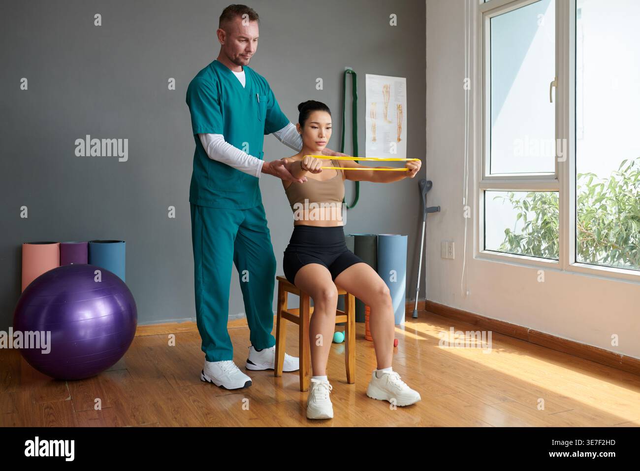 Asian female using stretch resistance band doing therapy assisted by caucasian male therapist sitting on chair in clinic Stock Photo