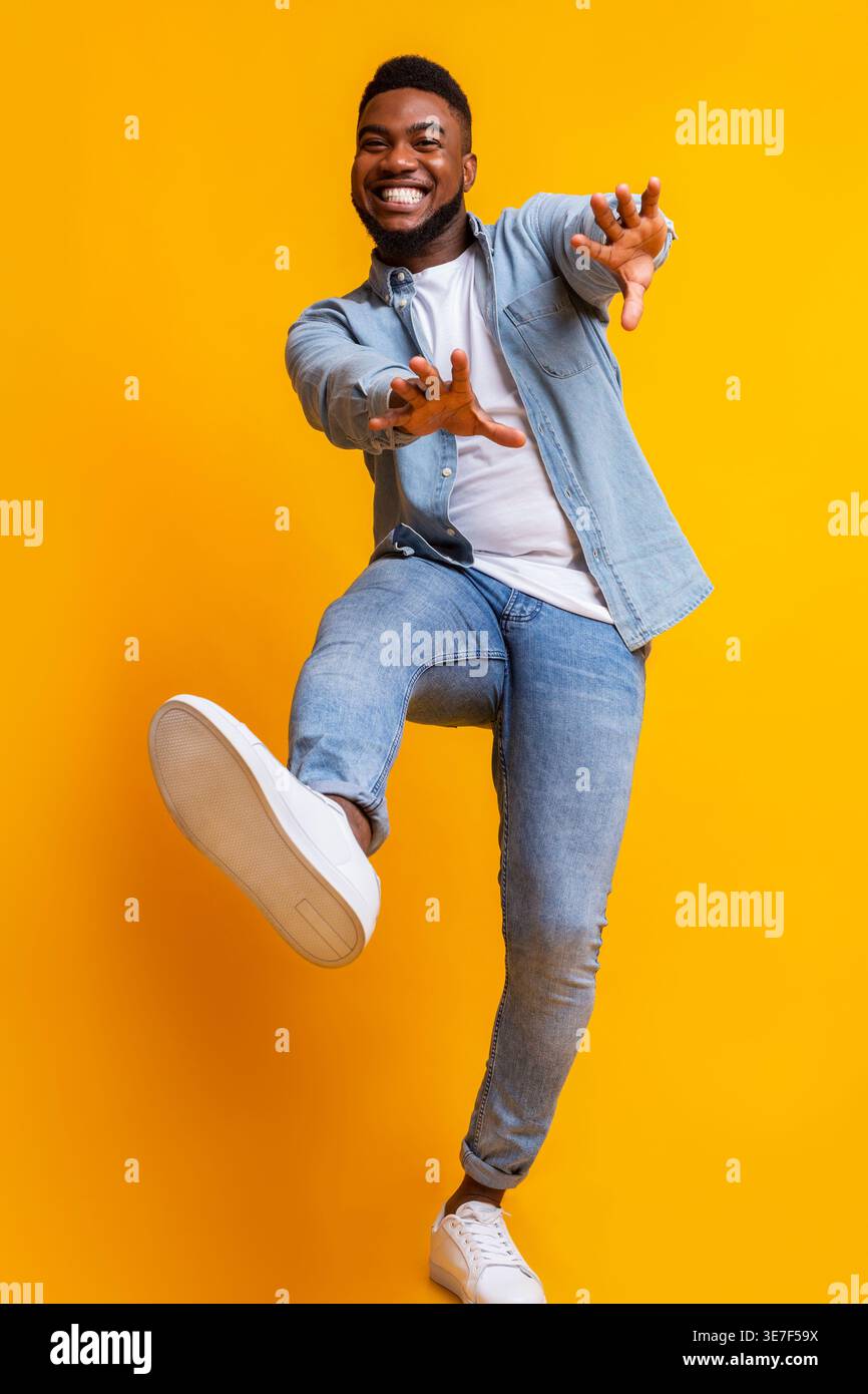 Cheerful Black man dancing and extending his hands forward against yellow background in studio Stock Photo