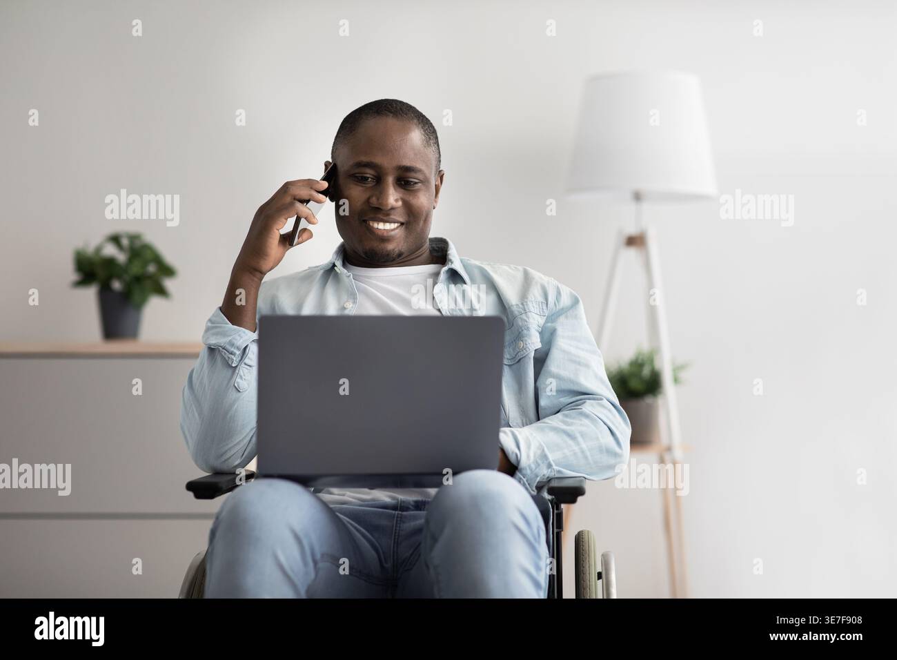 Disabled Black man sitting in wheelchair having phone call and looking at laptop screen working in living room at home Stock Photo