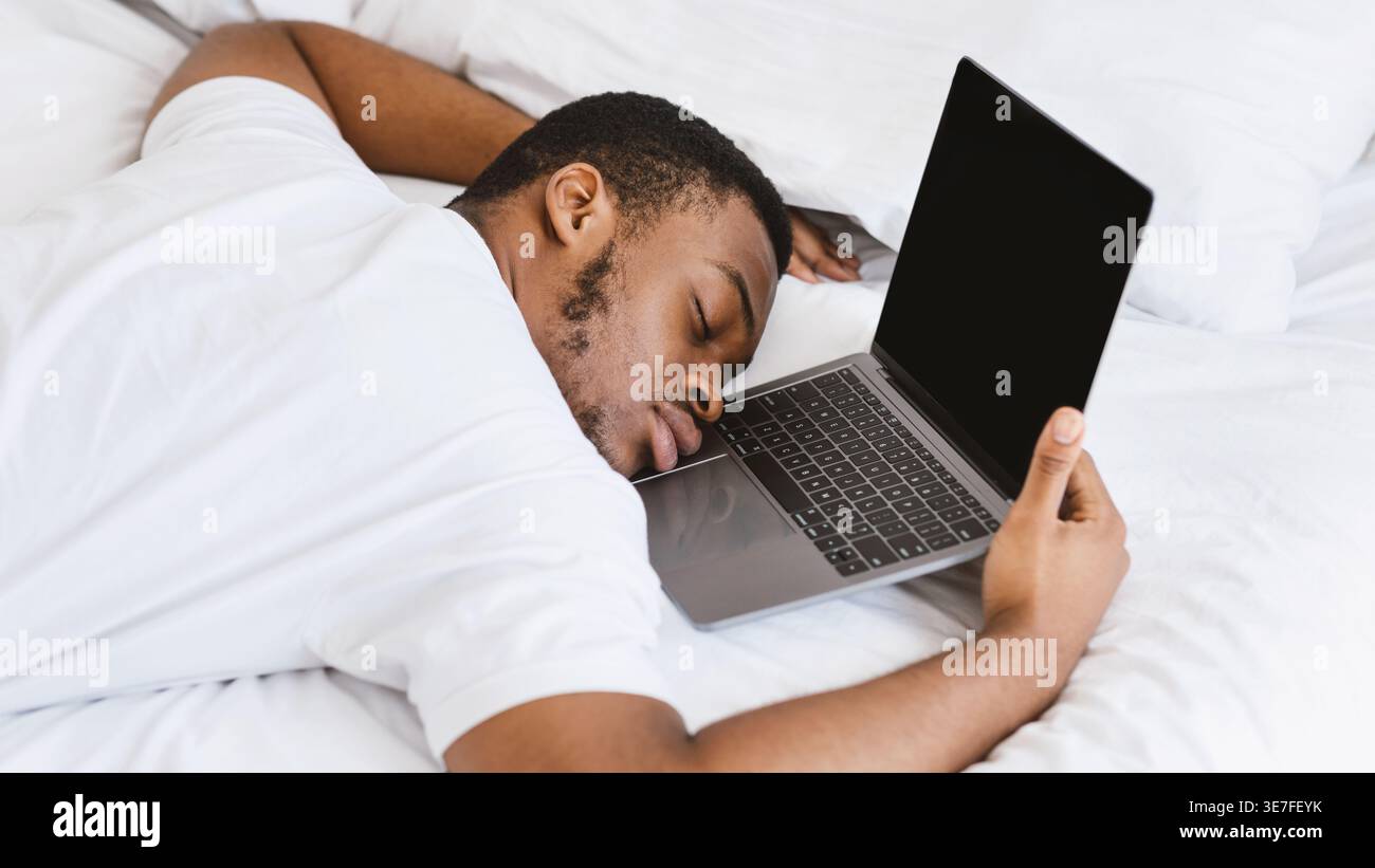 Black man in white shirt fell asleep while using laptop on bed at home Stock Photo