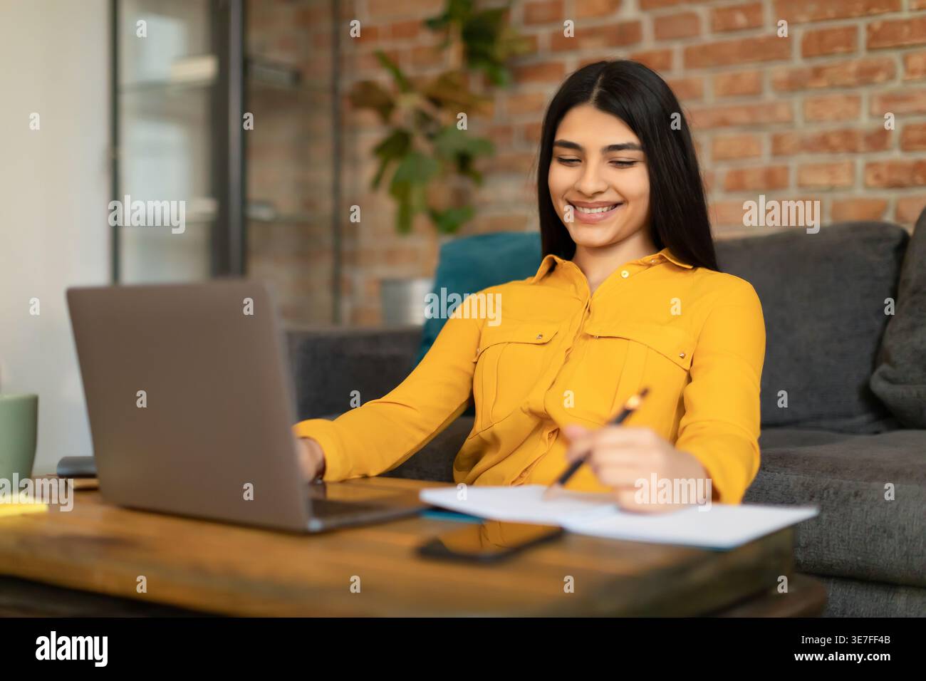 Middle Eastern female student using laptop while writing down notes sitting at desk in living room at home Stock Photo