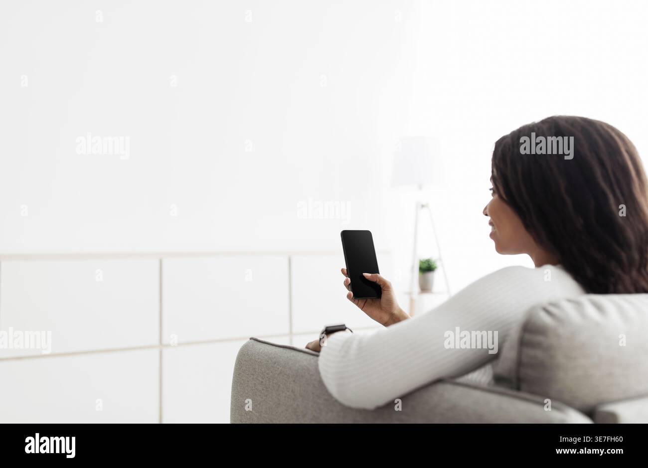 Black woman using smartphone sitting on sofa in living room at home Stock Photo