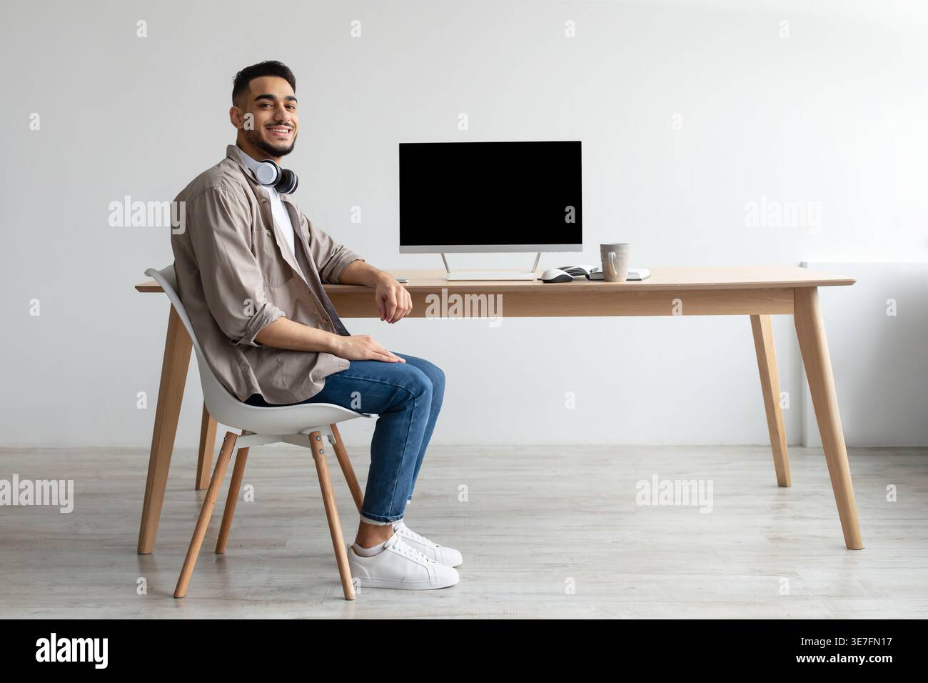 Smiling Middle Eastern man with headphones on neck sitting at table with computer and coffee cup in office Stock Photo