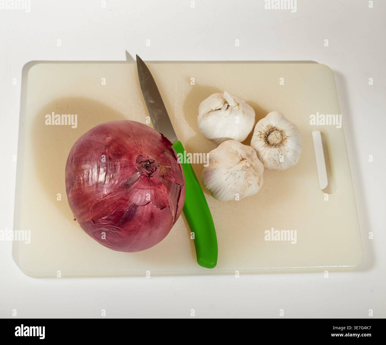 A red onion and three cloves of garlic on a cutting board with a green handled paring knife! Stock Photo