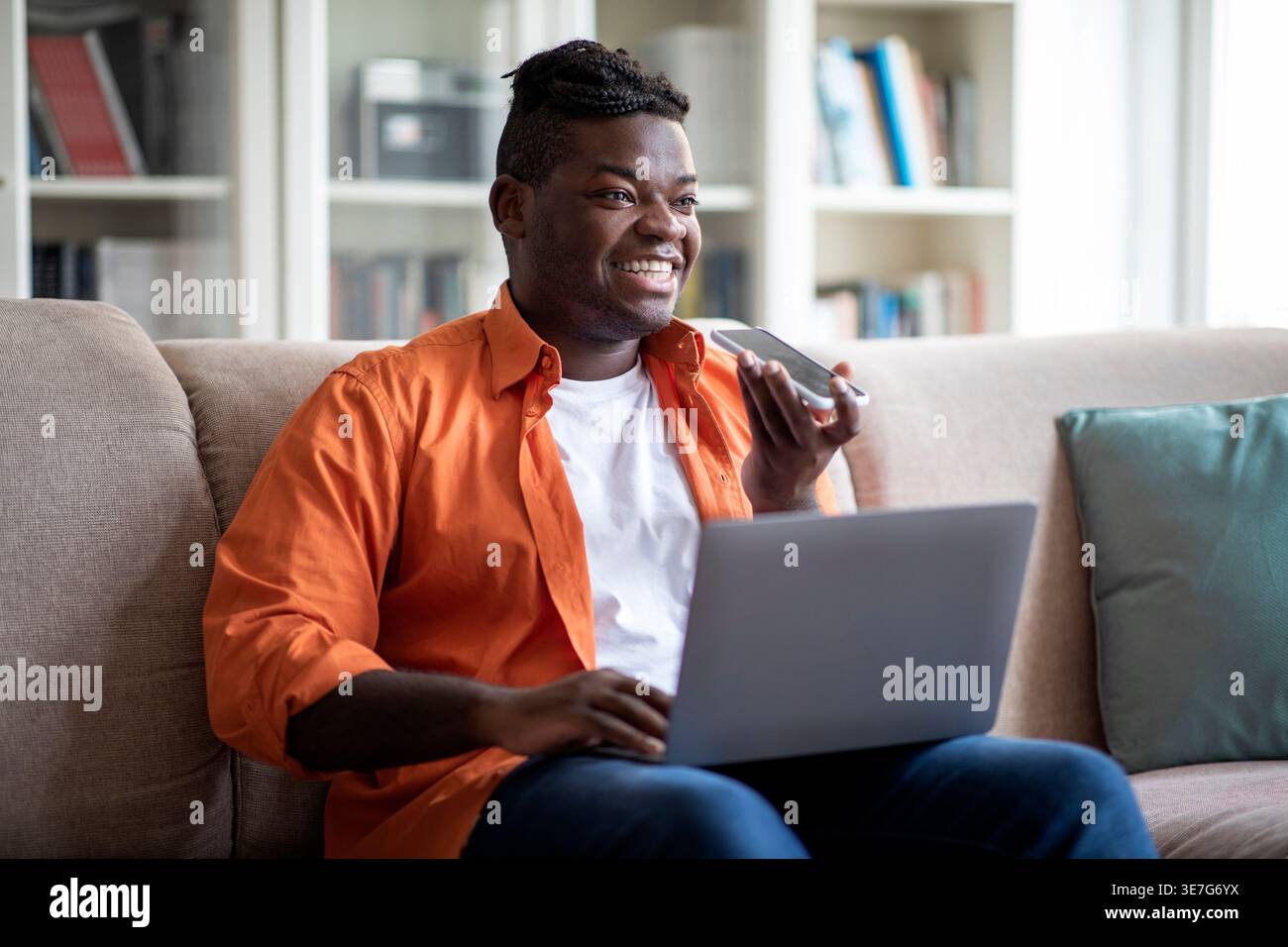 Smiling Black man with braids recording voice messages while using laptop sitting on sofa in living room at home Stock Photo