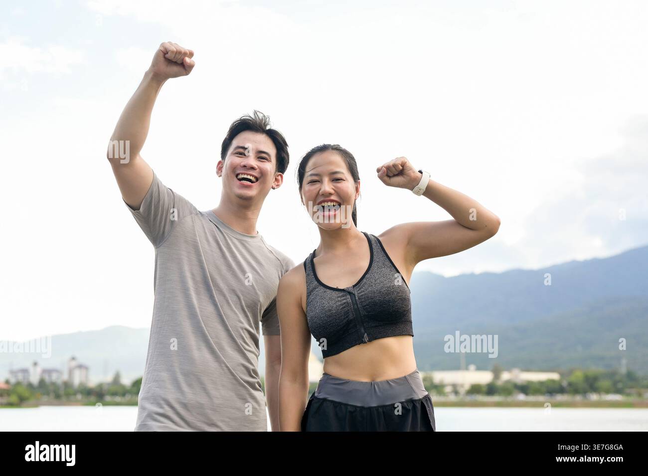 Cheerful Thai couple with raised arms and closed fists celebrating success after reaching their running target while standing together near lake in... Stock Photo