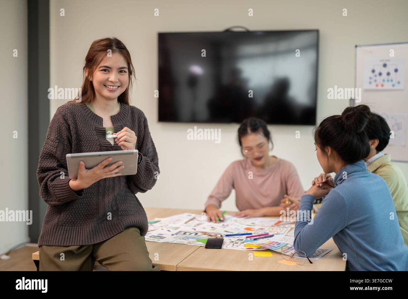 Smiling Asian woman holding tablet sitting on table near workmates in office Stock Photo