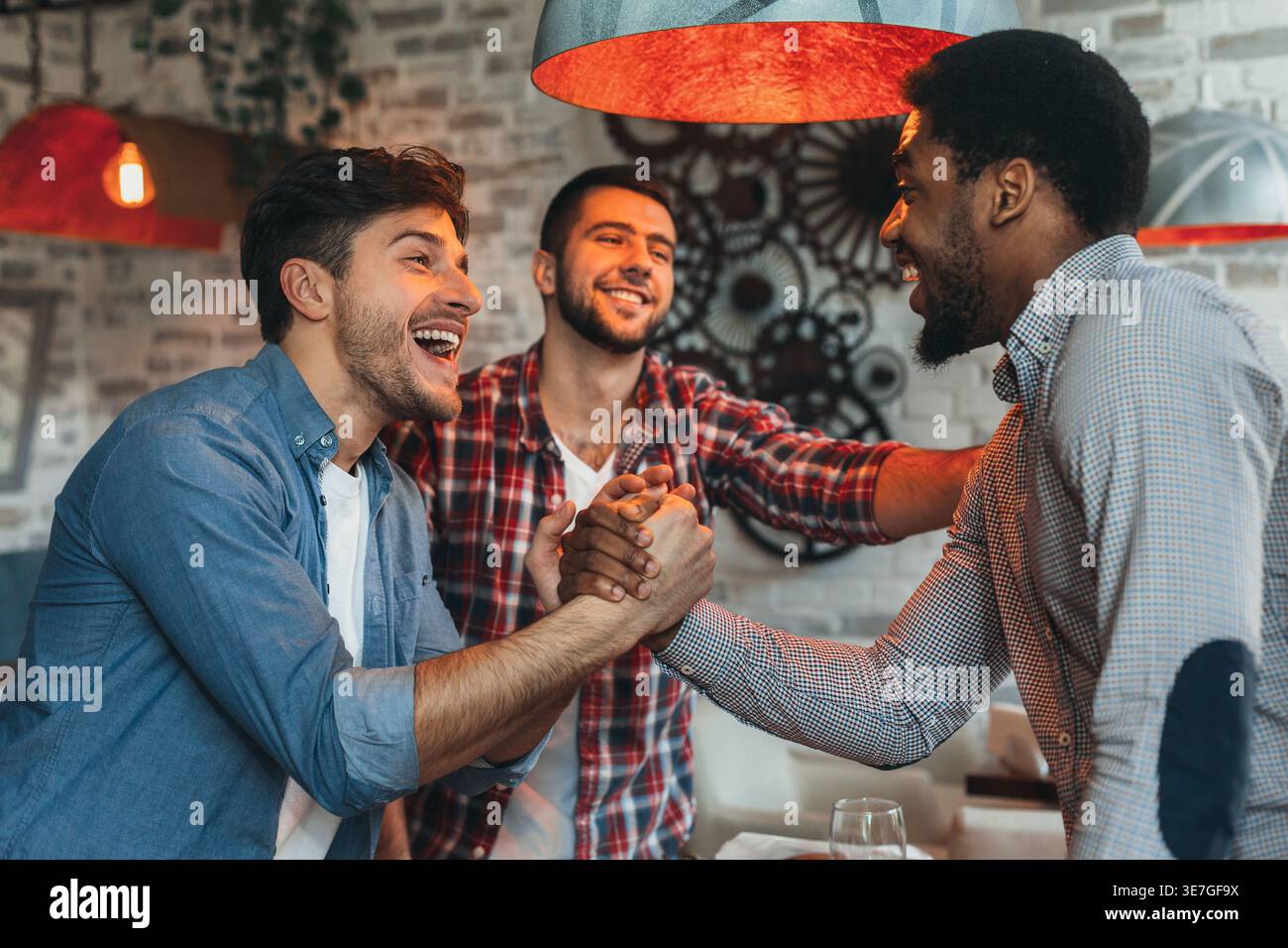 Cheerful Black man having fun and doing manly handshake with caucasian friends standing in bar Stock Photo