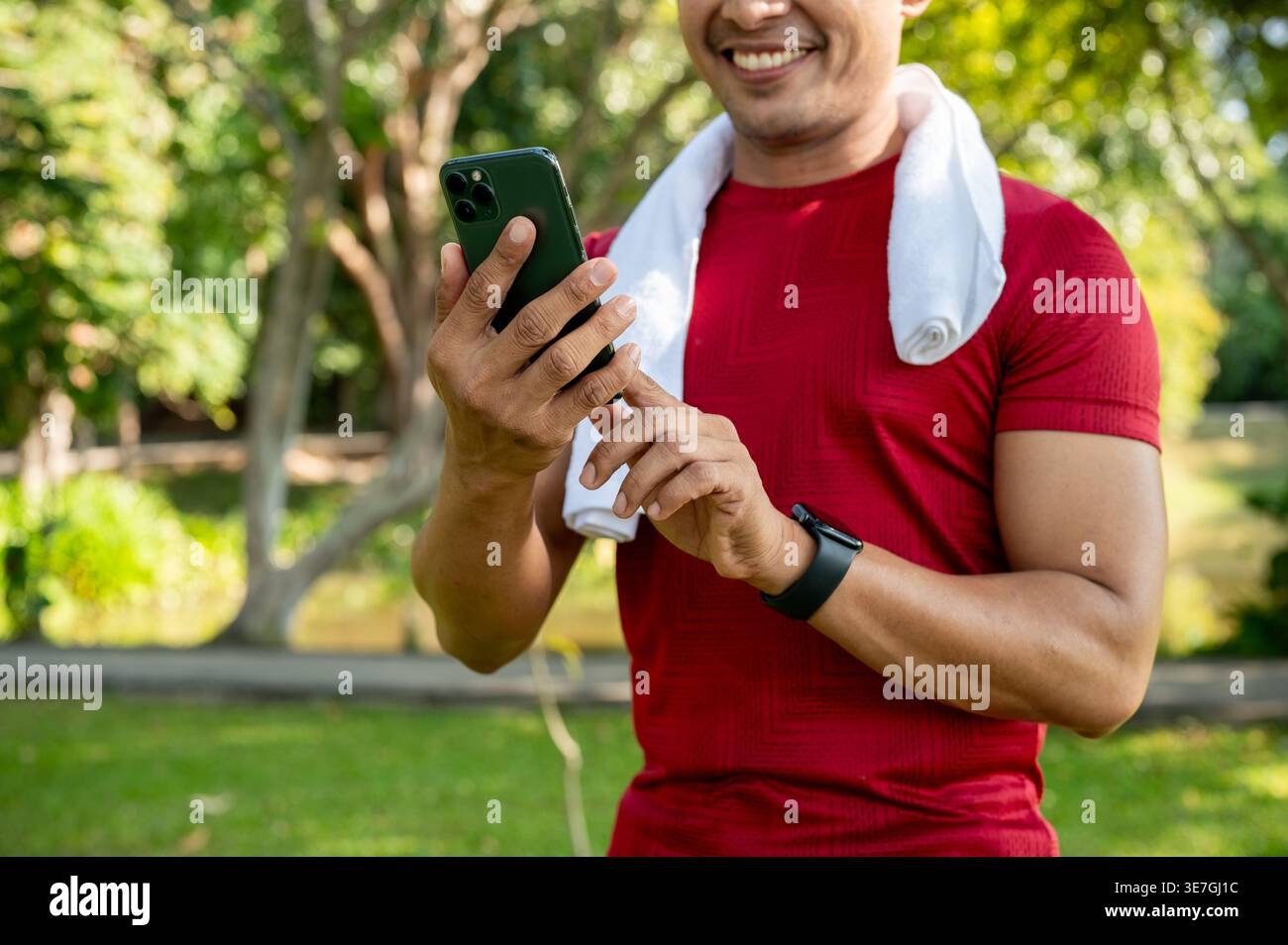 Cropped image of Thai man in fitness wear using smartphone while standing with white towel around his neck in park on sunny day Stock Photo