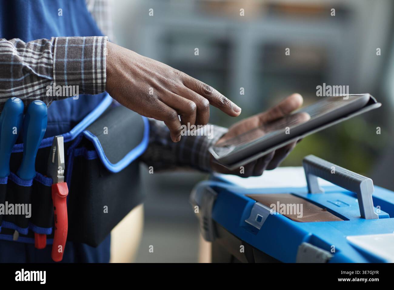 Cropped image of Black handyman with tool belt using tablet standing near toolbox indoors Stock Photo