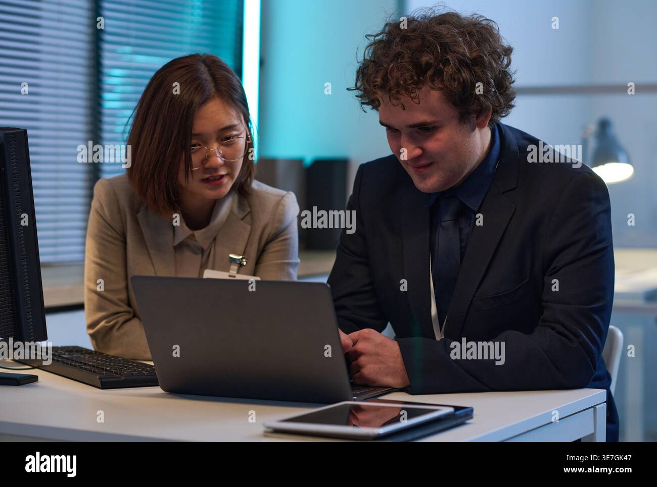 Asian female and caucasian male programmers sitting at desk discussing computer program on laptop working late at night in office Stock Photo