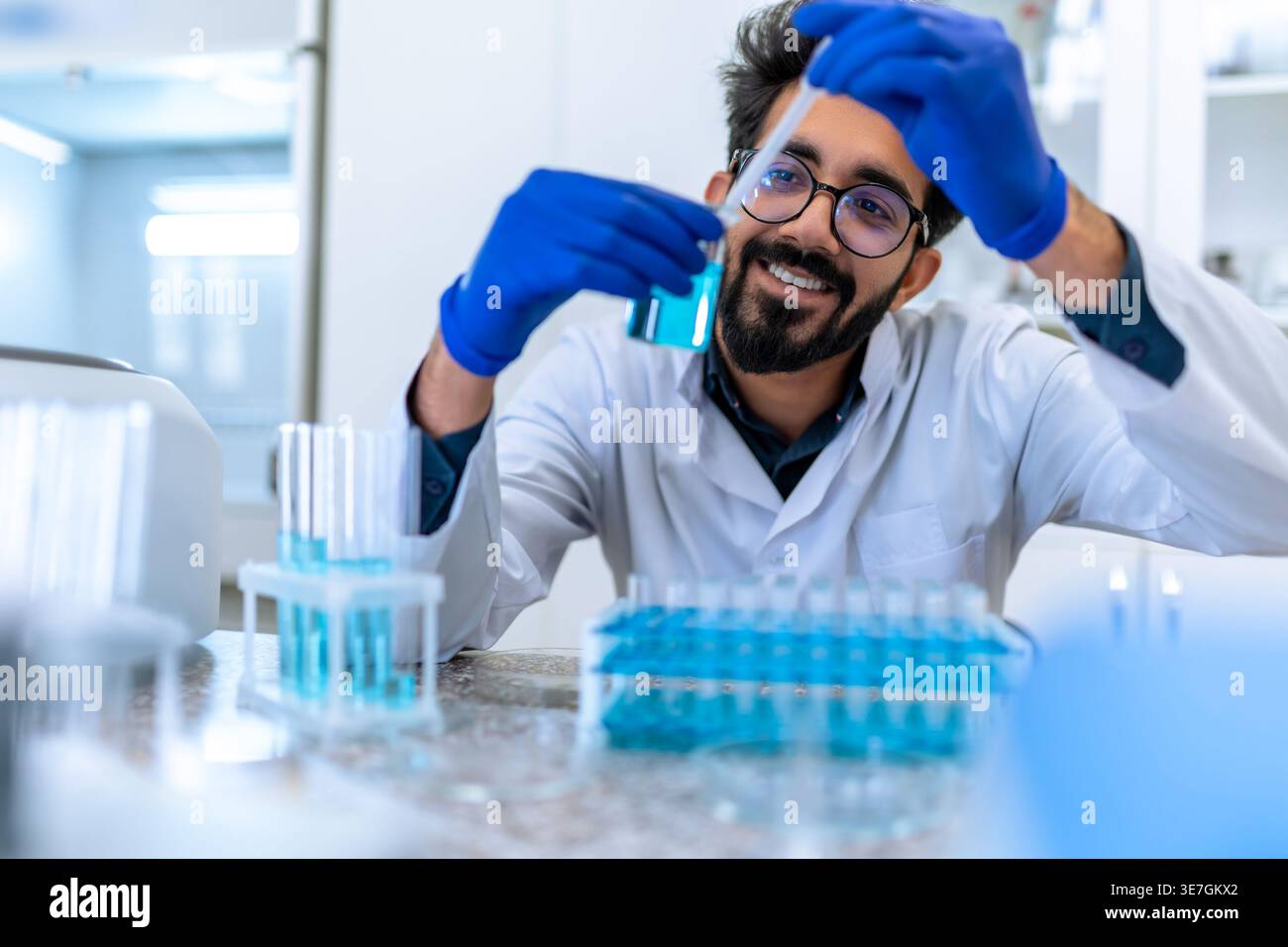 Smiling Indian male scientist wearing lab coat and latex gloves sitting at table and testing chemical sample in flask in laboratory Stock Photo