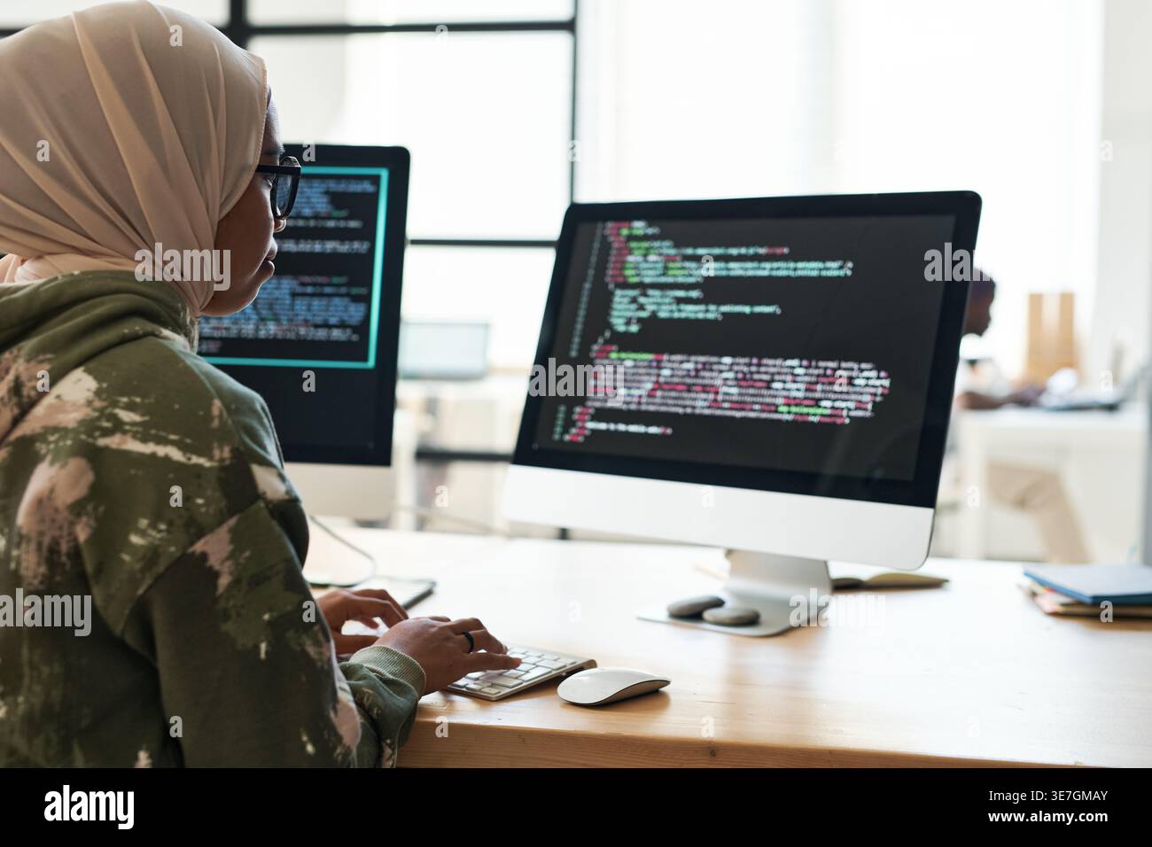 Focused Mixed-race Muslim female software developer decoding data working on computer with multiple screens sitting at table in office Stock Photo