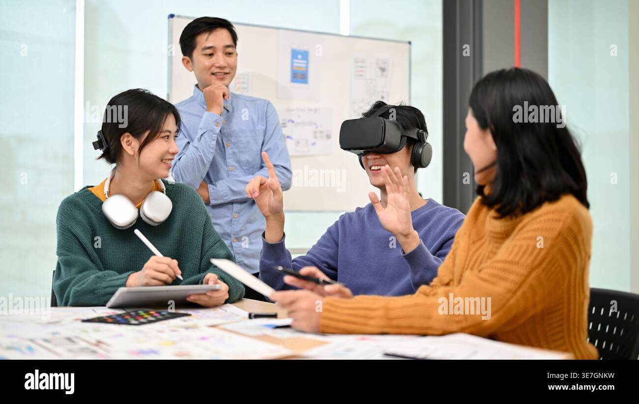 Smiling Asian male developer wearing VR headset sitting at table and testing newly developed VR game with colleagues in office Stock Photo