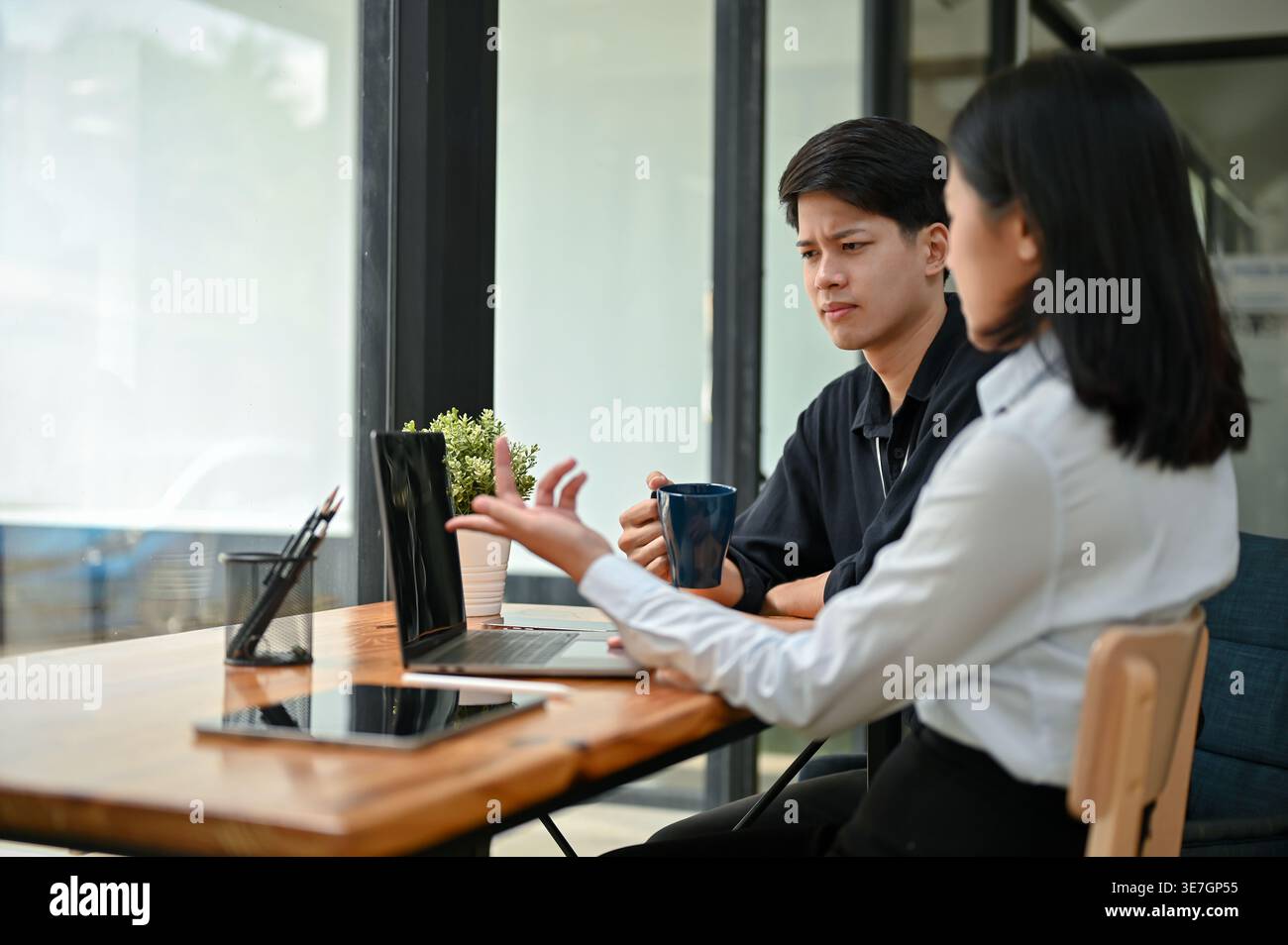Confused Asian male and female office workers pointing at laptop screen having problem with their project while sitting together at table in cafe Stock Photo
