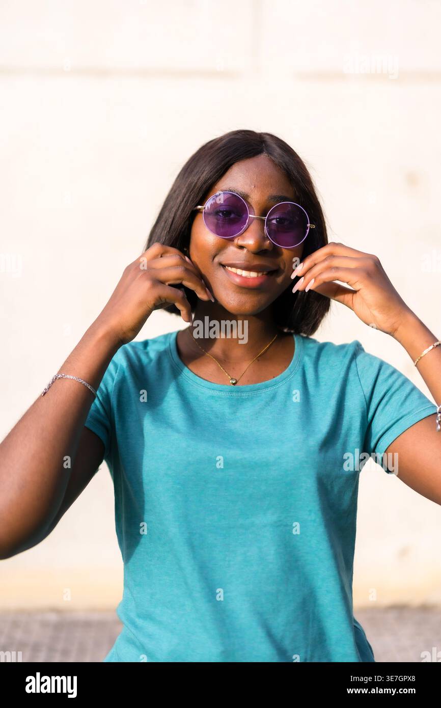 Smiling Black female in sunglasses touching face while standing against white wall Stock Photo