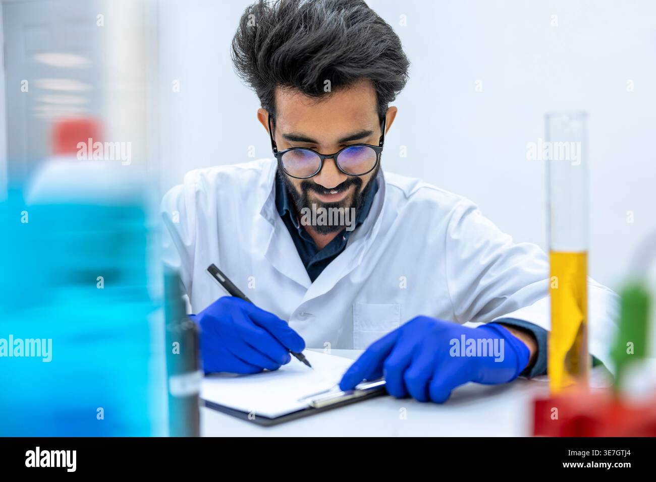 Smiling Indian male scientist wearing lab coat and latex gloves sitting at desk writing notes in laboratory Stock Photo