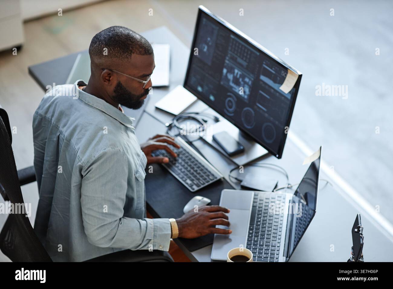 Black male programmer wearing eyeglasses using laptop while sitting at table with computer in office Stock Photo