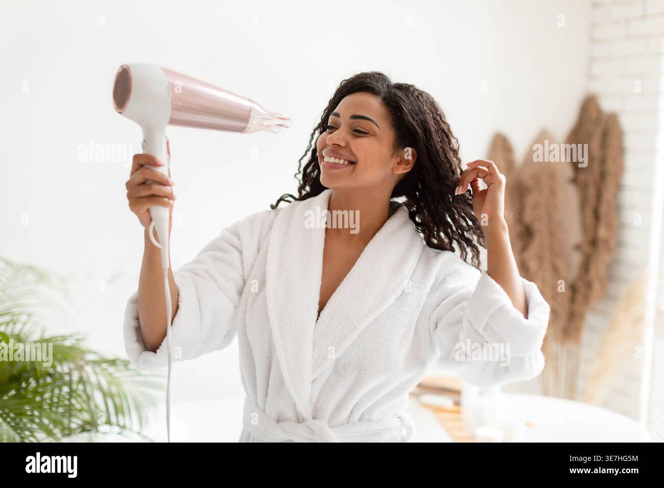Black woman in bathrobe blow drying hair standing in bathroom at home Stock Photo