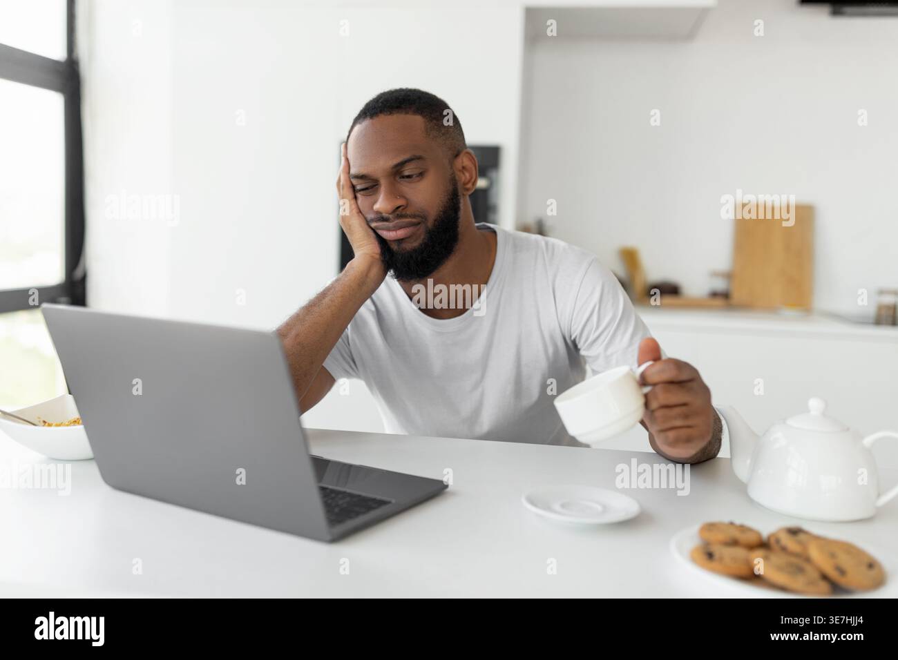 Bored Black man leaning face on hand holding coffee cup sitting at counter with laptop at home Stock Photo
