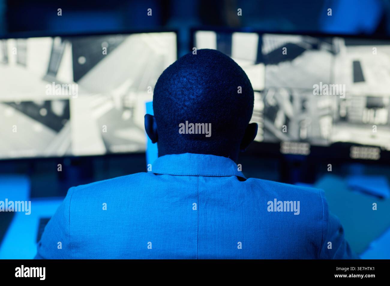 Back view of Black man looking at surveillance camera footage on computer while sitting at desk in security office at night Stock Photo