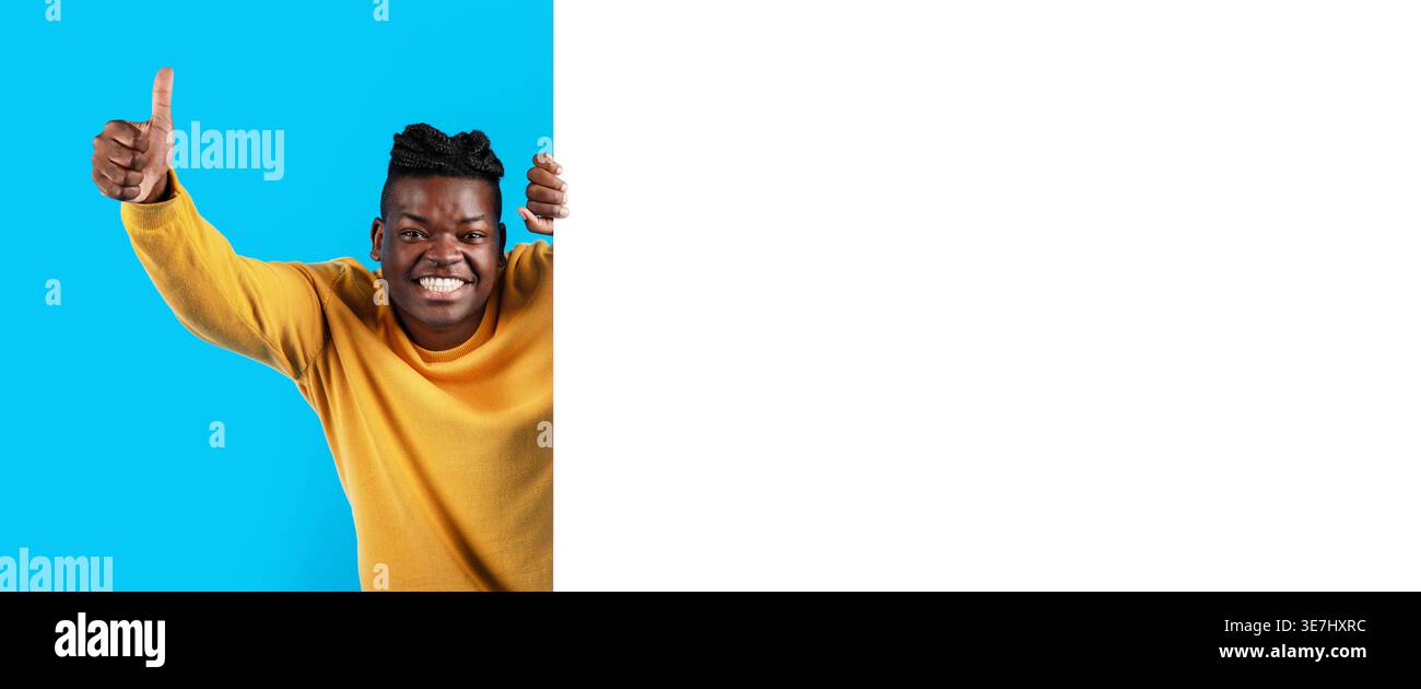 Cheerful Black man showing thumbs up hiding behind white blank advertisement board standing against blue background in studio Stock Photo