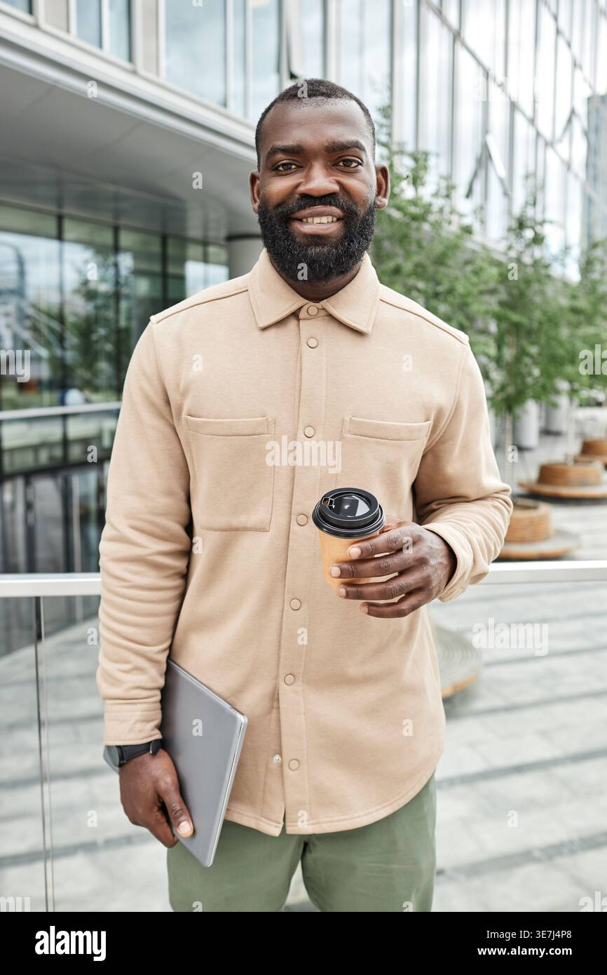 Black man holding laptop and takeaway coffee standing against glass fence in balcony on city during daytime Stock Photo