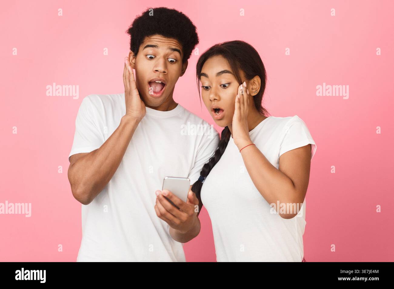 Shocked Black couple using smartphone while standing against pink background in studio Stock Photo