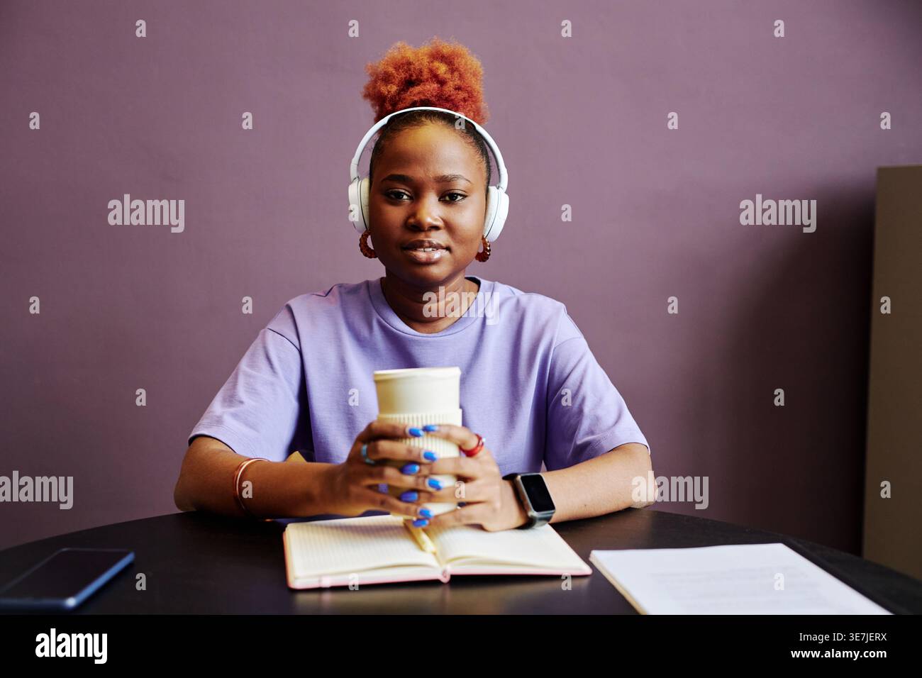 Black female programmer wearing wireless headphone holding coffee cup while sitting at table with notebook against purple background in studio Stock Photo