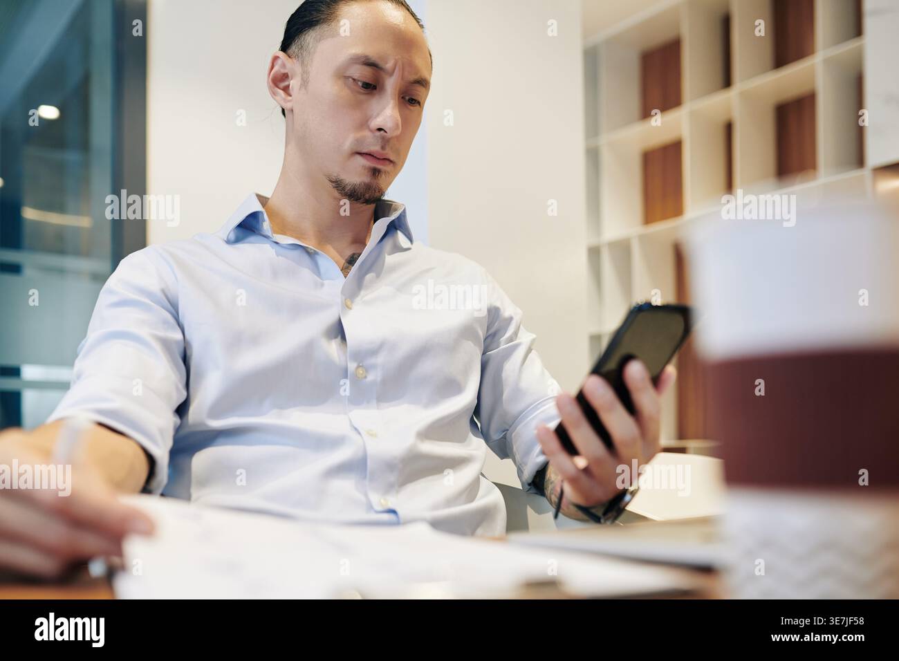 Frowning mixed-race businessman holding documents using smartphone sitting at table in office Stock Photo
