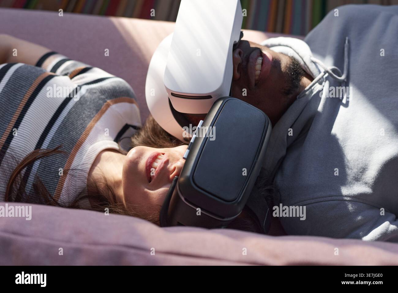 Black and mixed-race couple wearing VR headset lying on sofa together in living room at home Stock Photo
