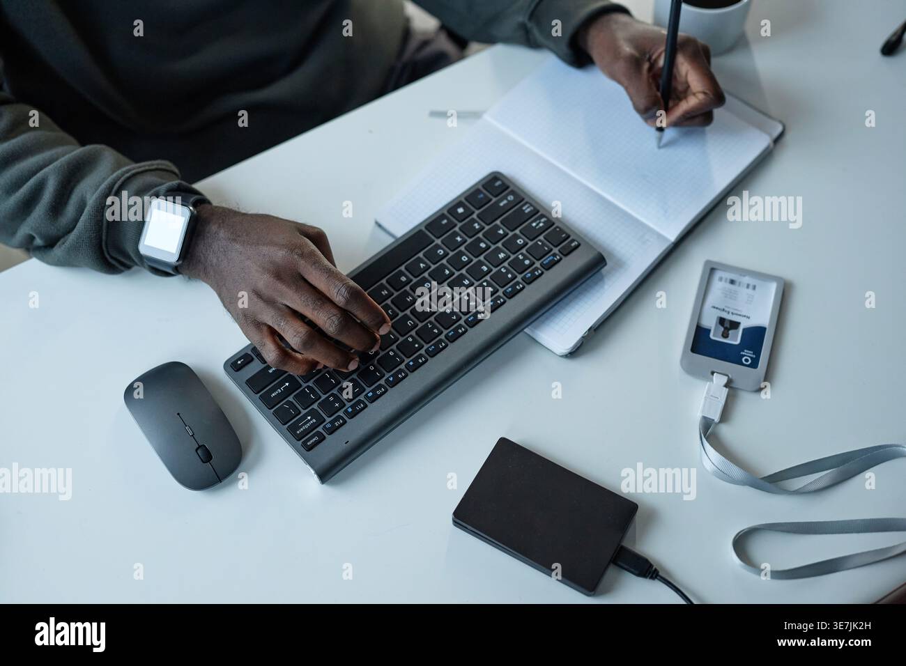 Hands of Black male programmer typing on computer using keyboard while taking notes sitting at table in office Stock Photo