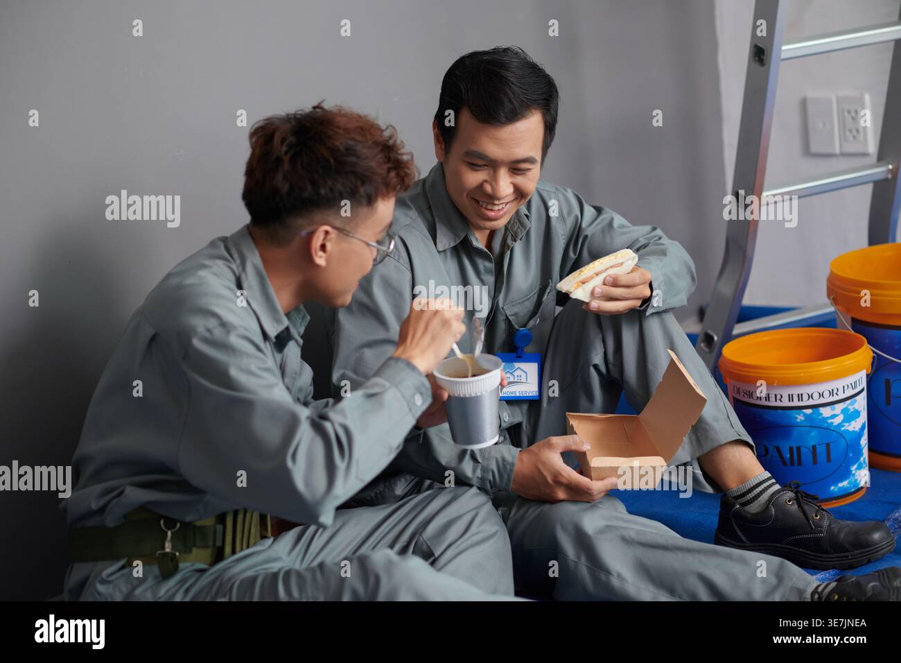 Asian male builders in uniform eating instant noodles and sandwich chatting during lunch break sitting on floor against gray wall on construction site Stock Photo