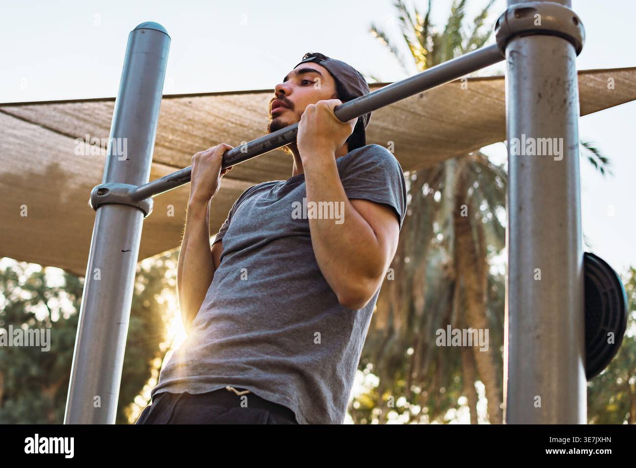 Middle Eastern man with beard wearing cap doing pull-ups in outdoor gym during sunny day Stock Photo