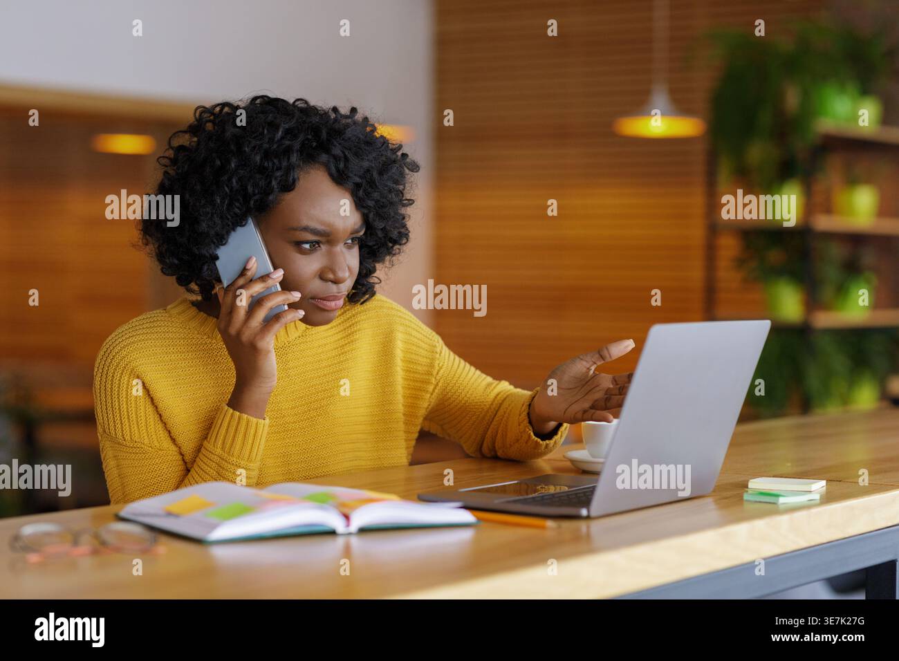 Serious Black woman with curly hair gesturing hand having phone call looking at laptop sitting at table with notebook in cafe Stock Photo