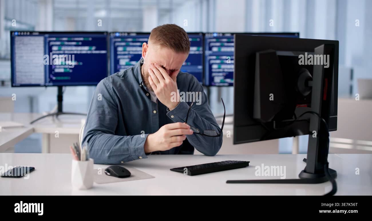 Tired Coder Rubs Irritated Eye During Extended Hours At Workstation Stock Photo