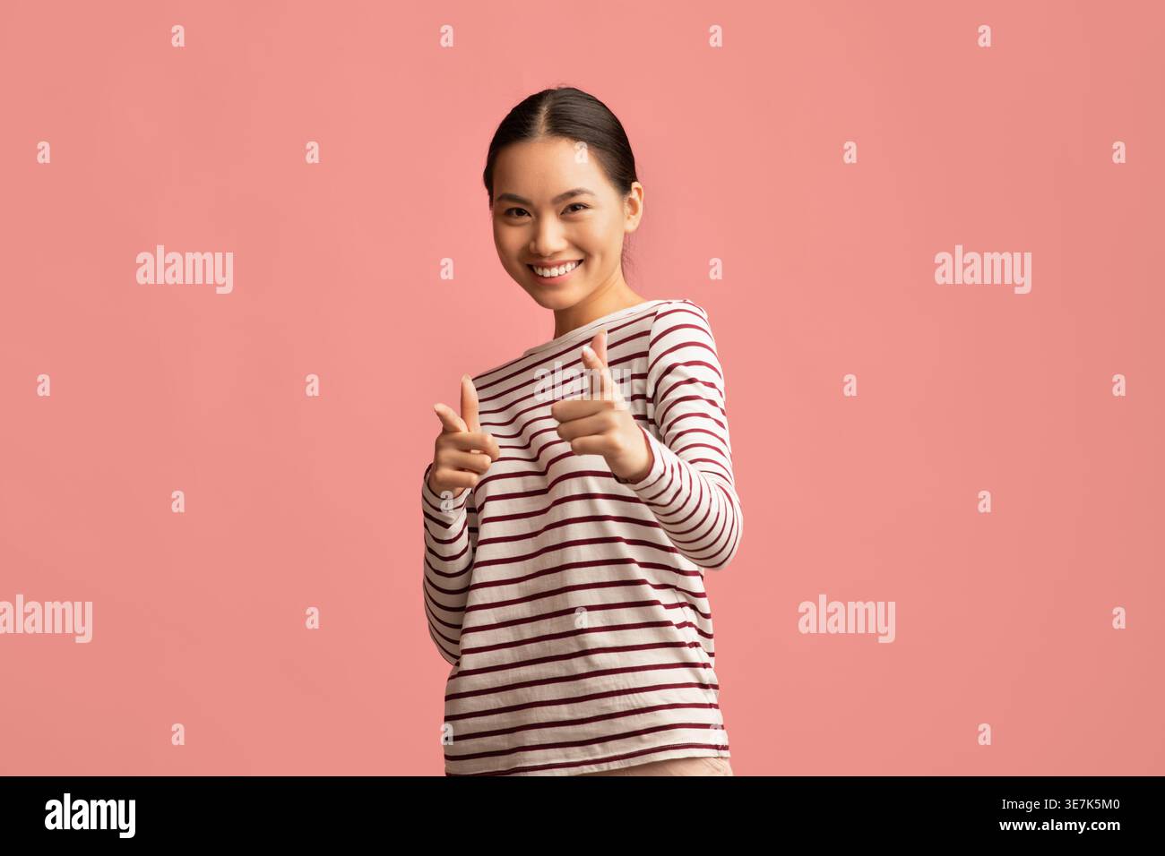 Smiling Asian female pointing fingers forward standing against pink background in studio Stock Photo