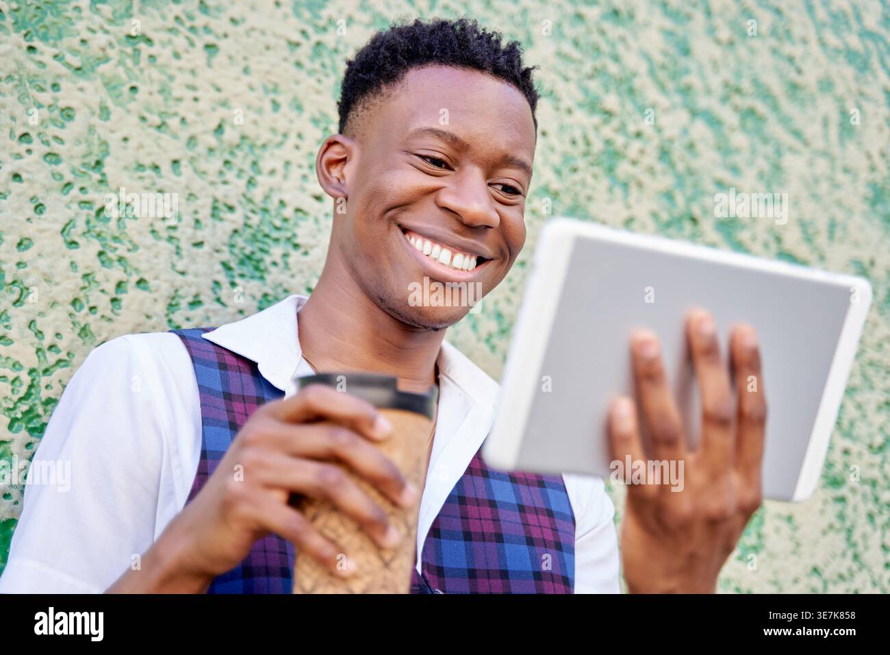 Black man holding takeaway coffee and using tablet while standing in front of concrete wall outdoors Stock Photo