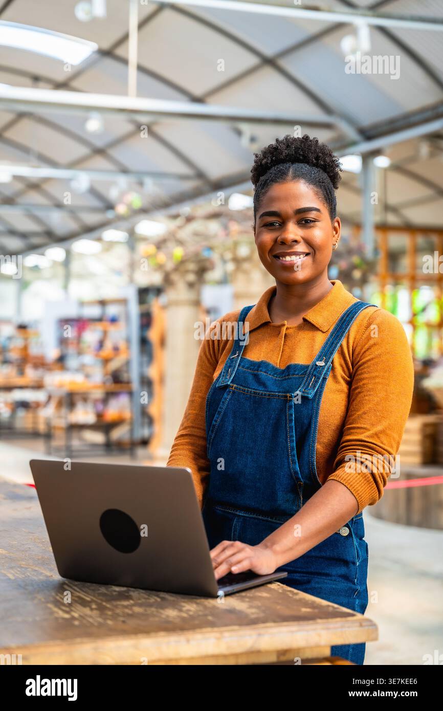 Hispanic female employee managing orders using laptop while standing at wooden table in garden center Stock Photo