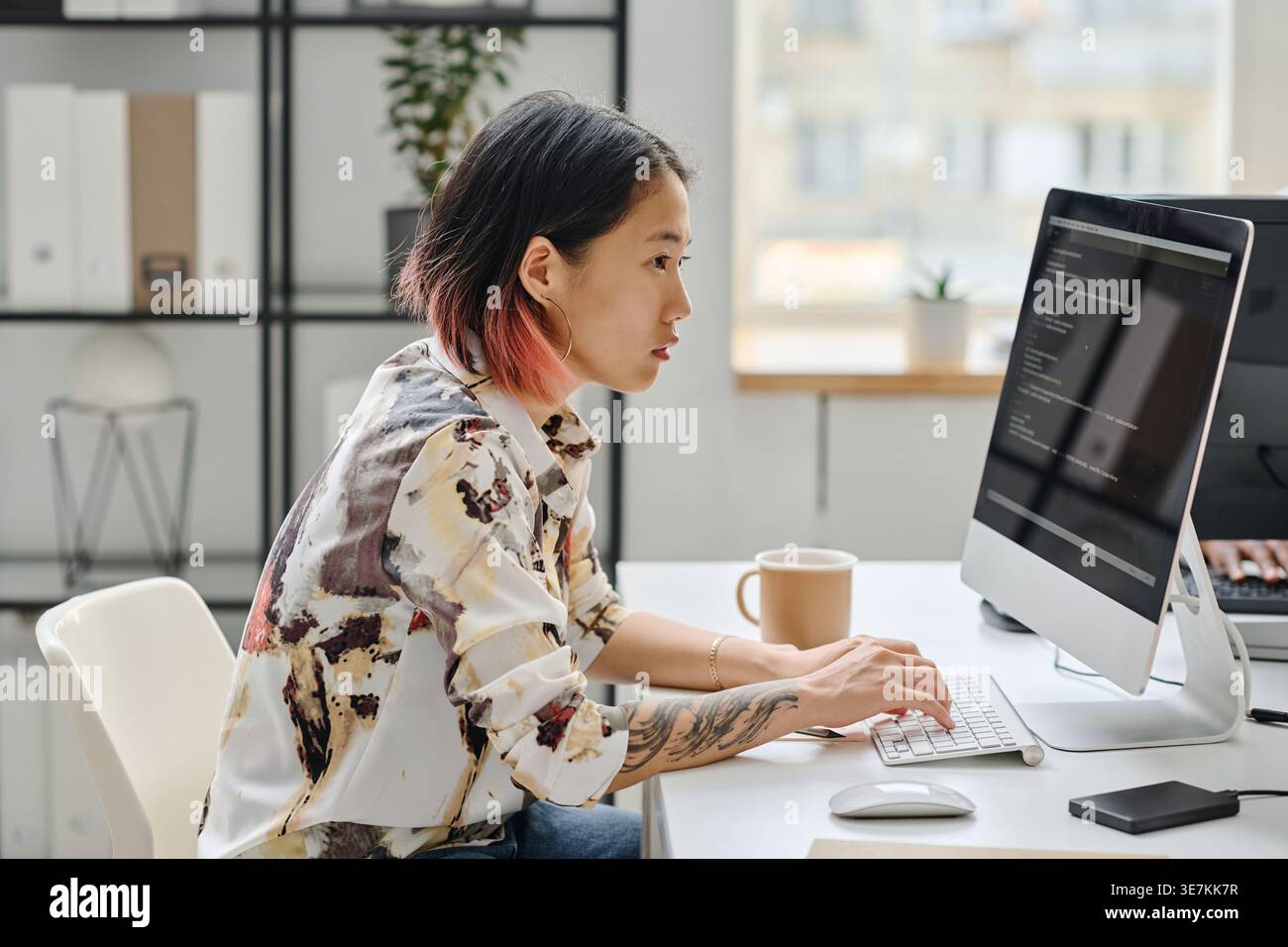 Asian female programmer working on computer typing codes while sitting at desk with coffee in office Stock Photo