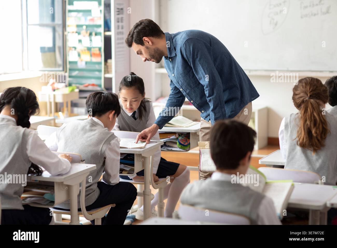 Diverse group of students doing school work while caucasian male teacher assisting them in classroom Stock Photo